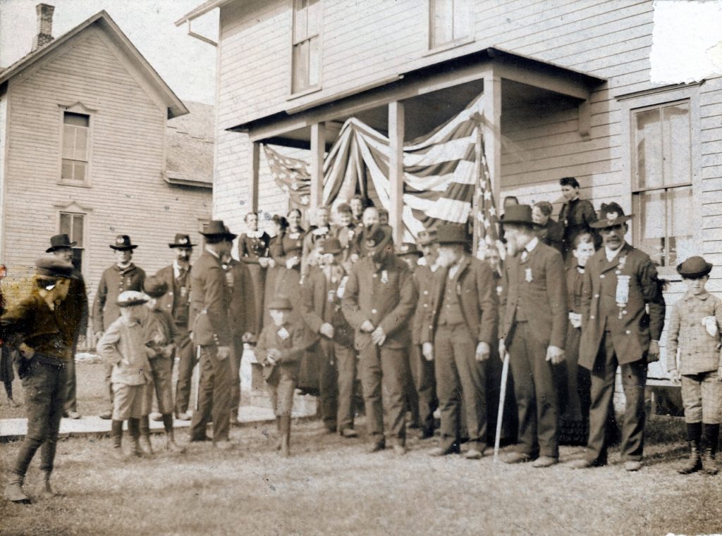 The above is a rare photo of Manistique’s Civil War veterans getting ready for a patriotic holiday celebration. Pictured from far right: John Gayar, Henry Brassel, Amos Hill, George W. Rice  (partially hidden behind Hill) and Wright E. Clarke (buttoning coat), On the far left, the men with insignias on their hats are David Blair (left) and William Wood (Right).