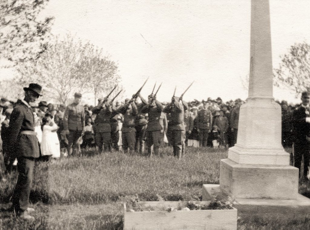 Early 1900s photo of a Decoration Day ceremony at the G.A.R./Spanish American War monument at Lakeview Cemetery. Civil War veteran Gear stands in front of the monument while Spanish American War veterans fire a salute. A box of flowers can be seen in the foreground, ready to be strewn upon the graves of the soldiers resting there. Niles and Helmka Family Collection.